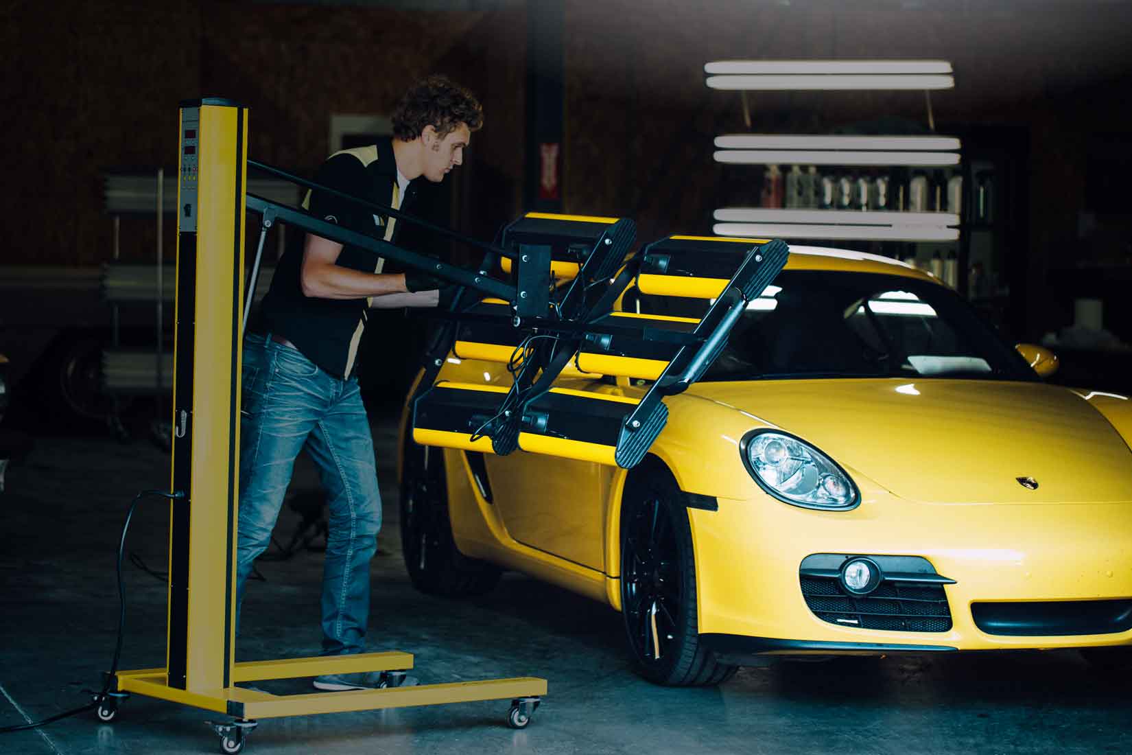 Un técnico de RestorFX apuntando una lámpara infrarroja en un auto deportivo amarillo durante la restauración en una superficie brillante y reluciente