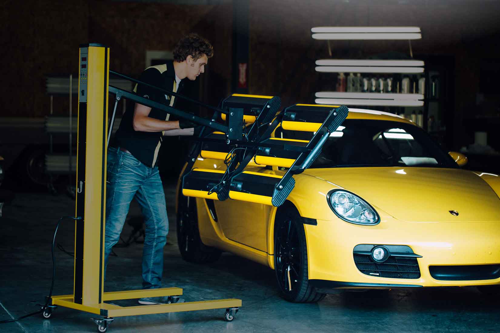 Un technicien RestorFX braque une lampe infrarouge sur une voiture de sport jaune pendant la restauration sur une surface lumineuse et brillante