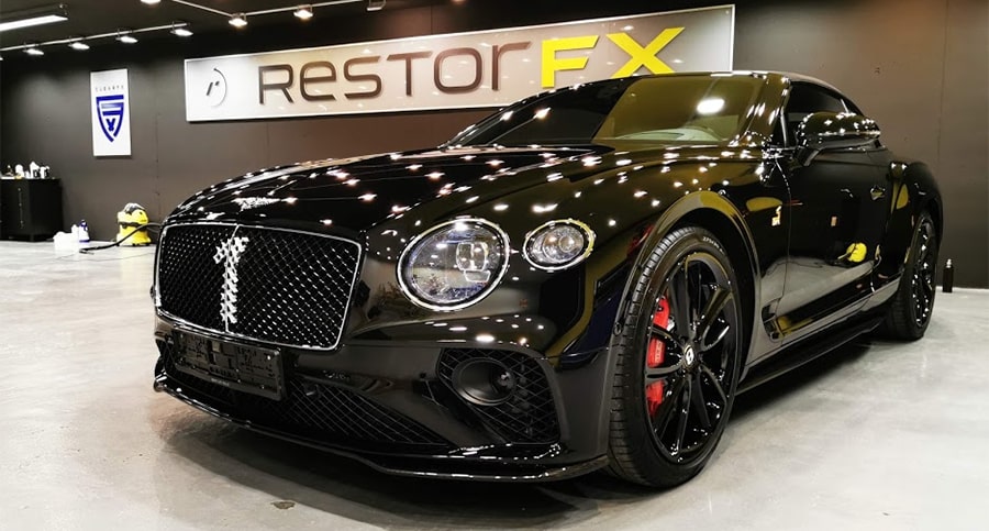 A lustrous black car in a bright RestorFX Center work area with black walls, yellow accent ceiling panel and lights