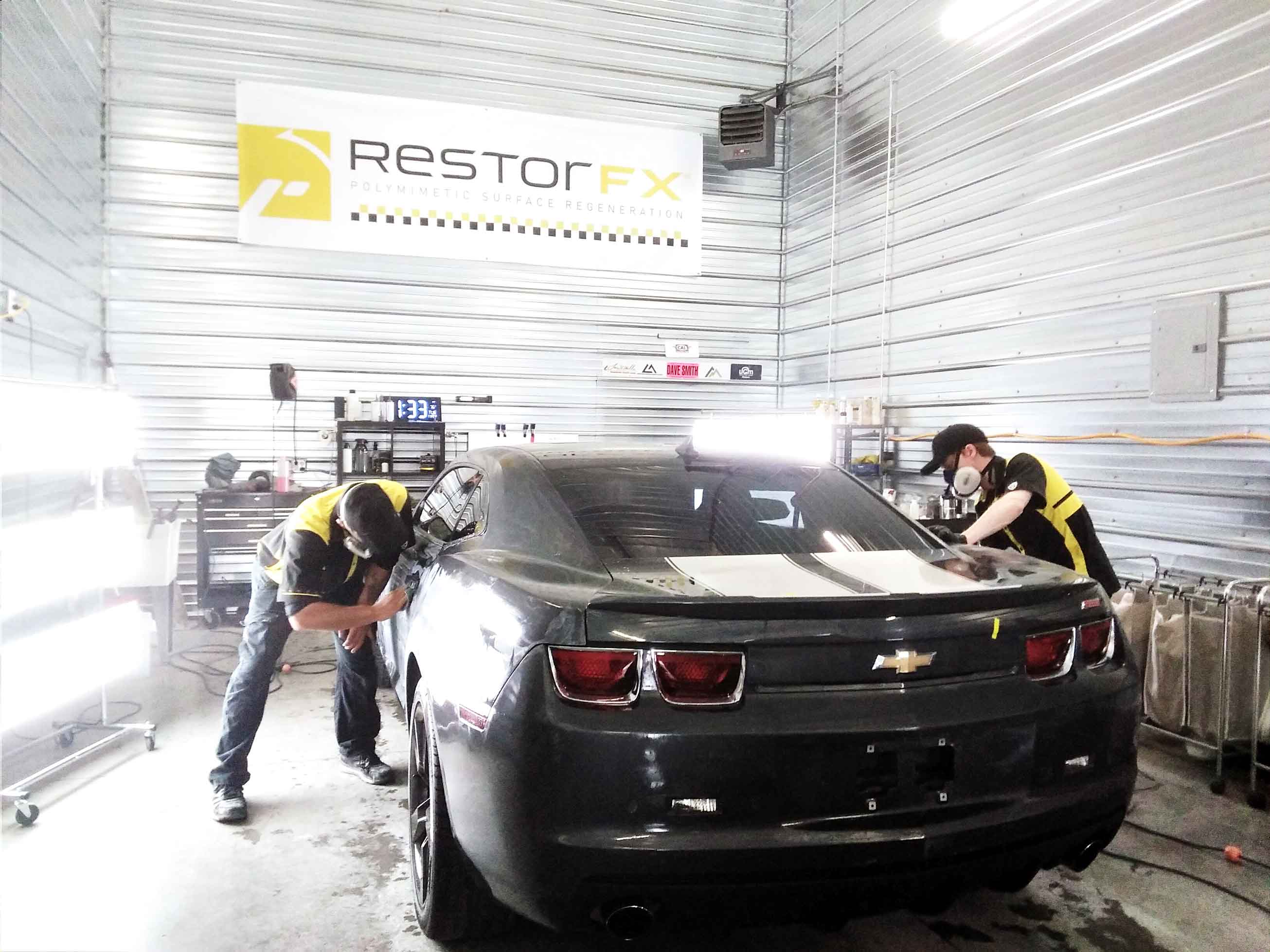 Technicians restoring black Camaro in brightly lit shop area at RestorFX Spokane with branded wall