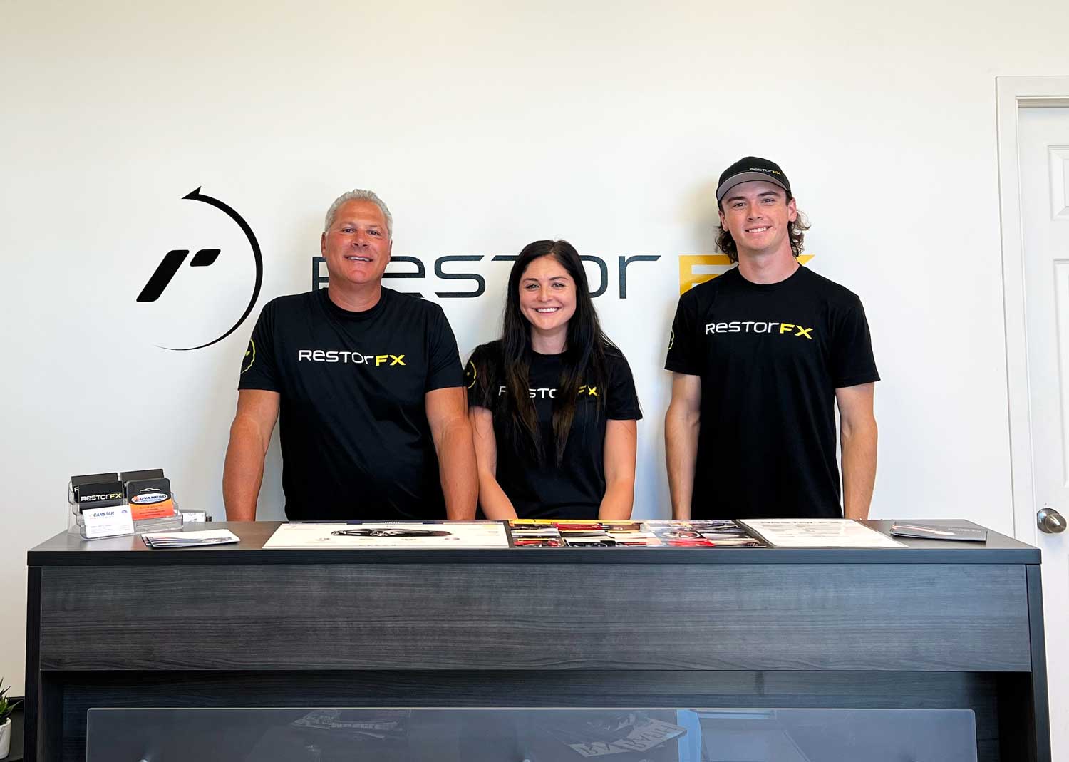 RestorFX Windsor team standing proudly in front of logo on branded white wall in the lobby area