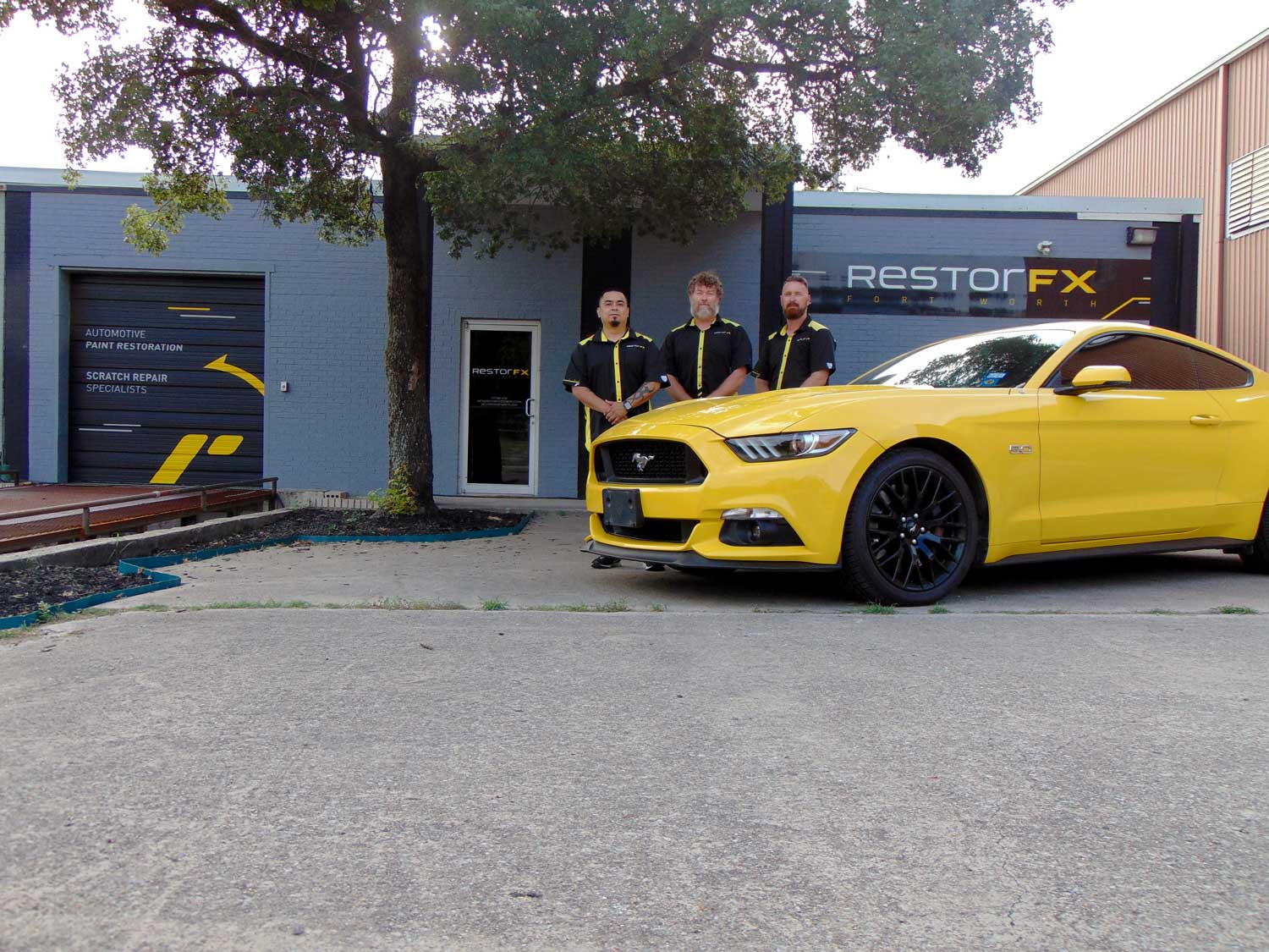 RestorFX Fort Worth team in front of the Center with a gray brick facade, branded signage and a yellow Ford Mustang sports car