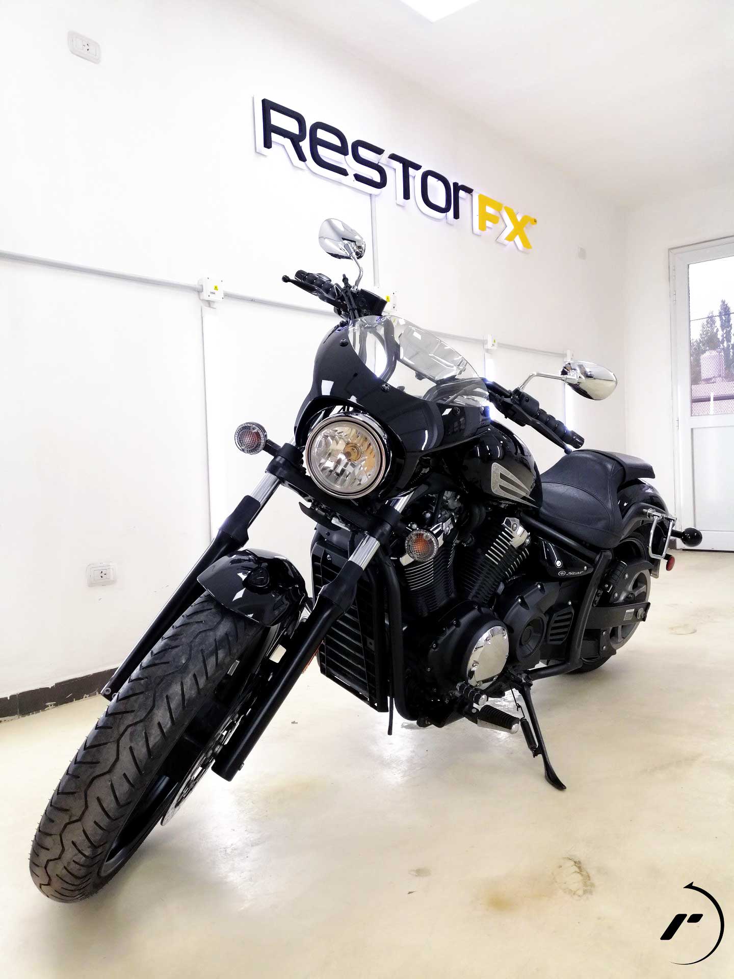 Shiny black motorcycle at RestorFX Comodoro with white wall, lights and branded signage in the back