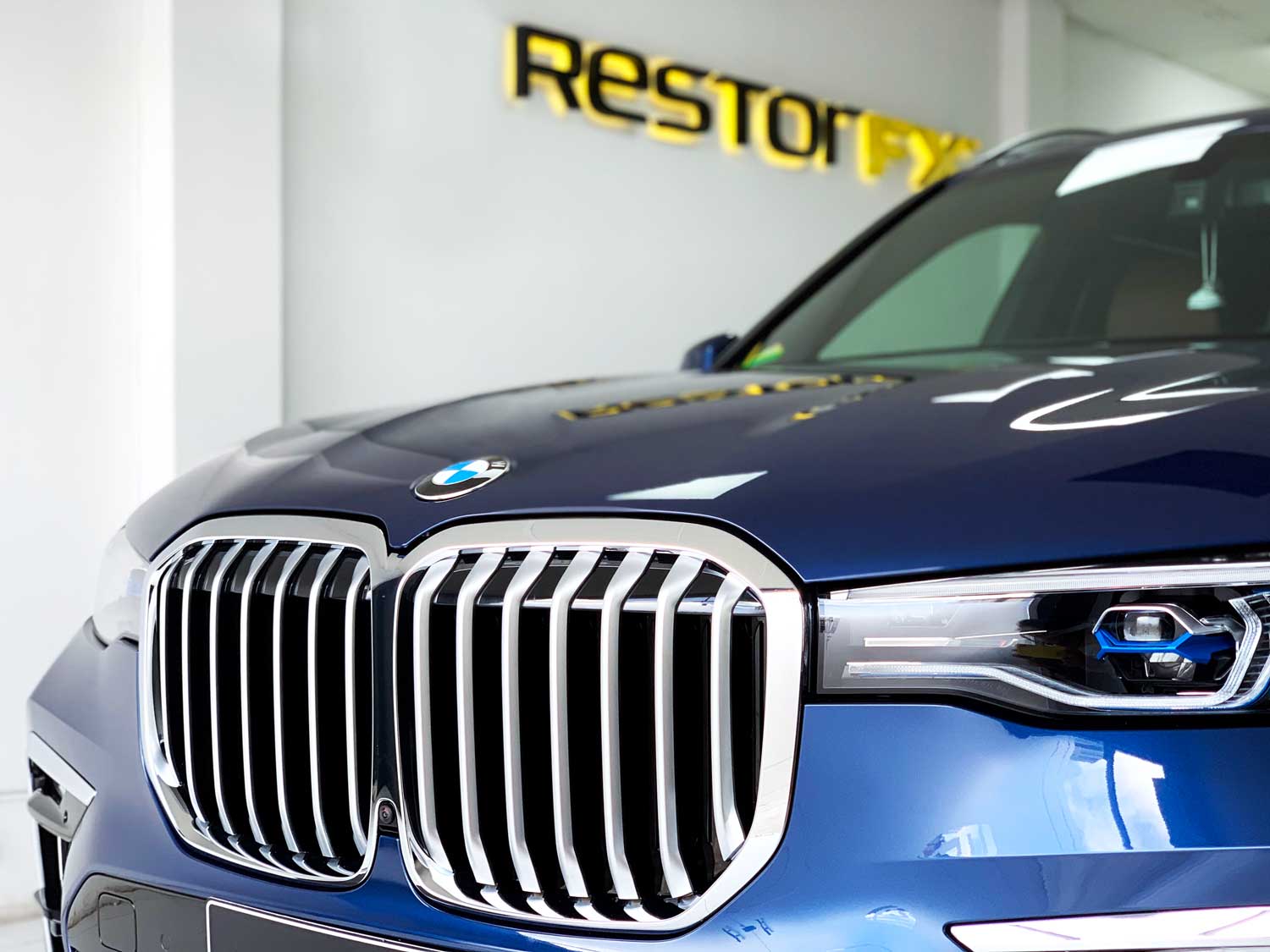 Angled-view of blue BMW with grille and headlight at the front and branded white wall at the back