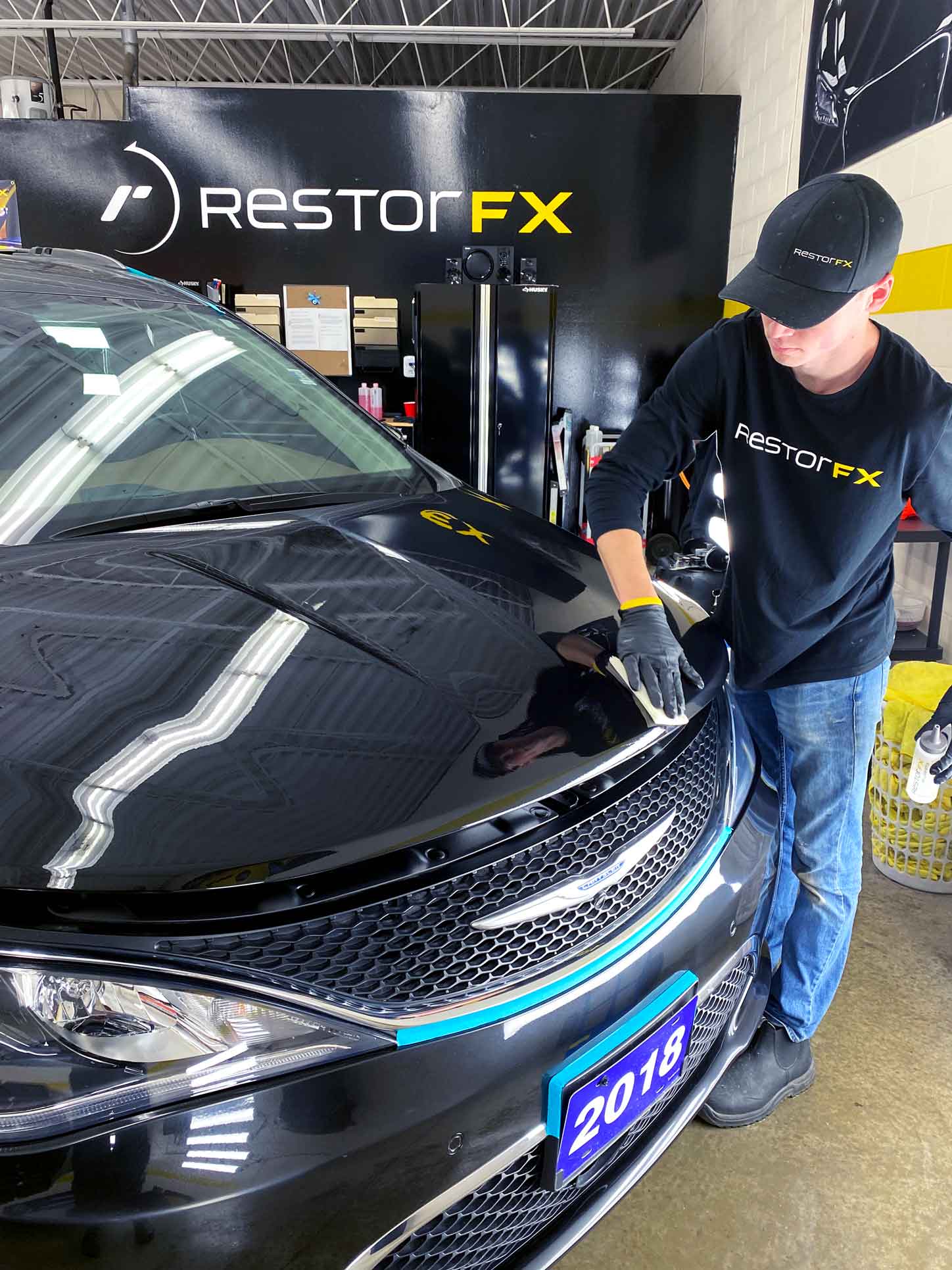 Technician restoring the front panel of a black Chrysler in branded shop area at RestorFX Windsor