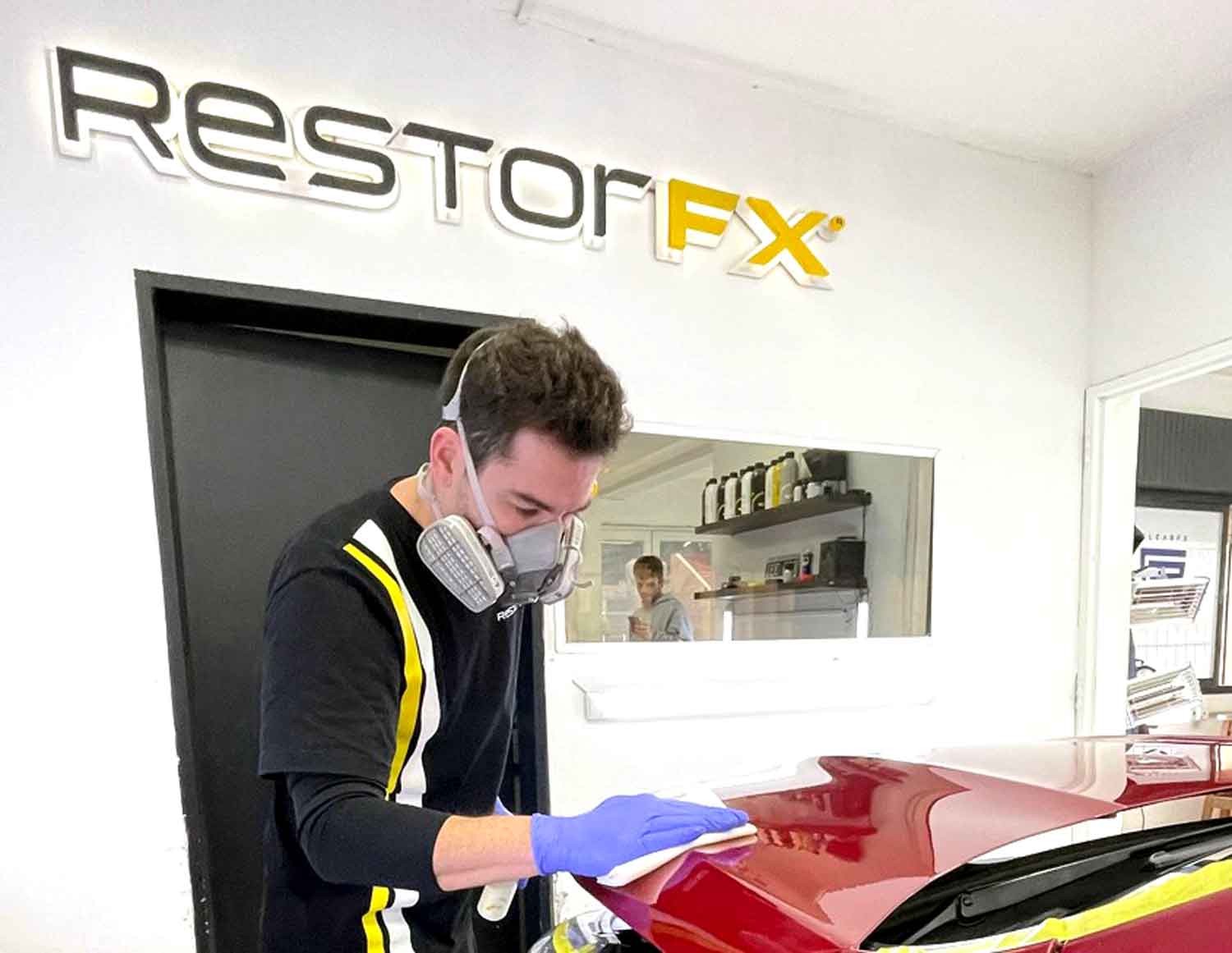 Technician applying RestorFX product onto red vehicle's surface during paint restoration process with branded sign in the backgroundTechnician applying RestorFX product onto red vehicle's surface during paint restoration process with branded sign in the b