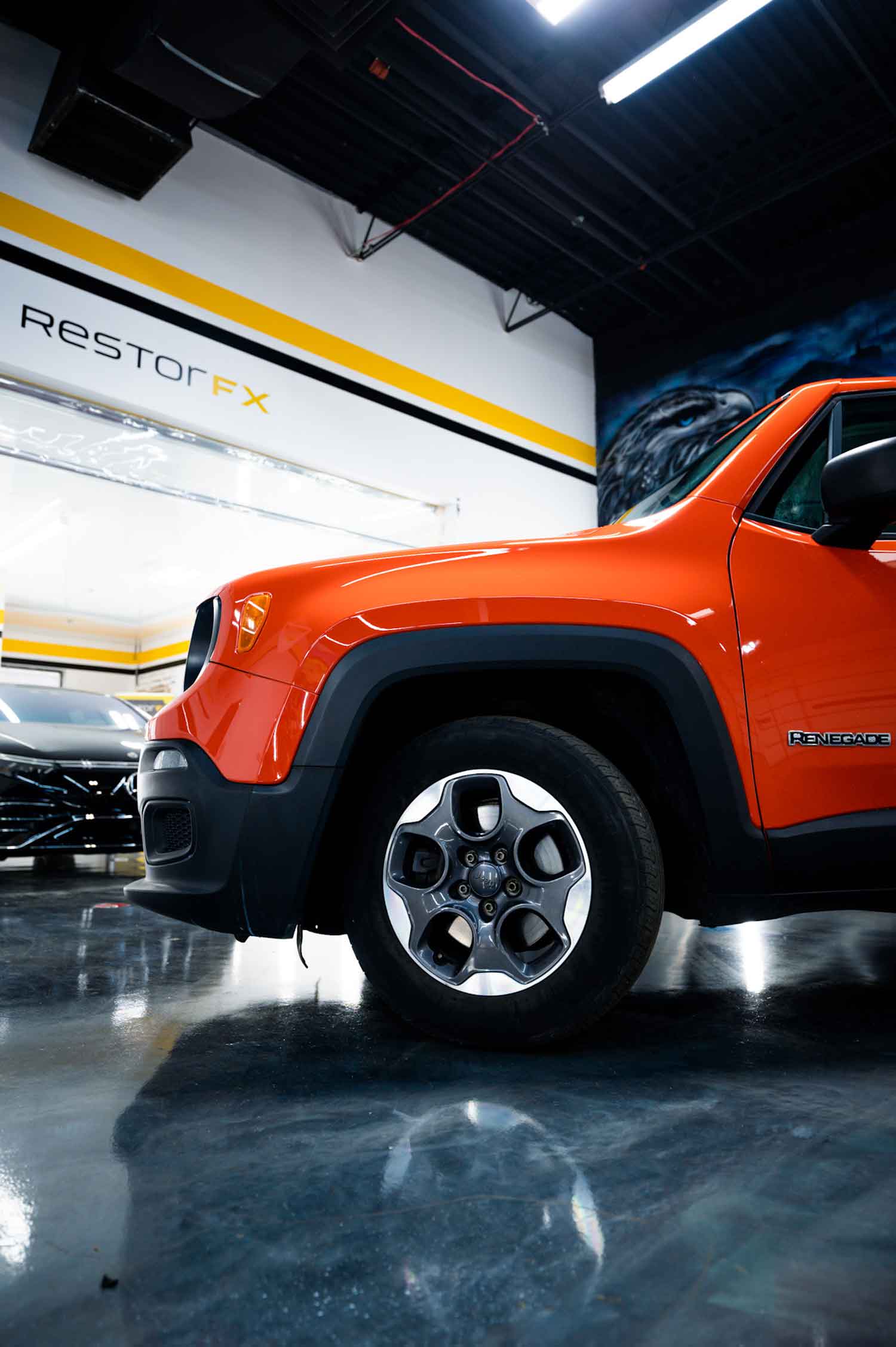 Glossy orange Jeep Renegade parked in brightly lit shop area at RestorFX Buffalo Grove with branded wall vinyls and black car in the background