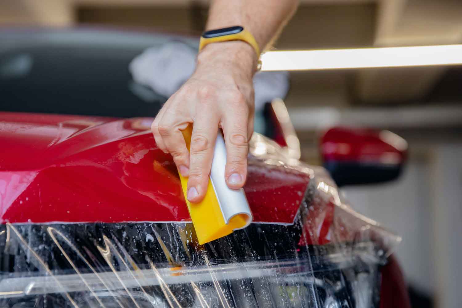 Technician applying clear protection film to the hood of a red car using a yellow squeegee