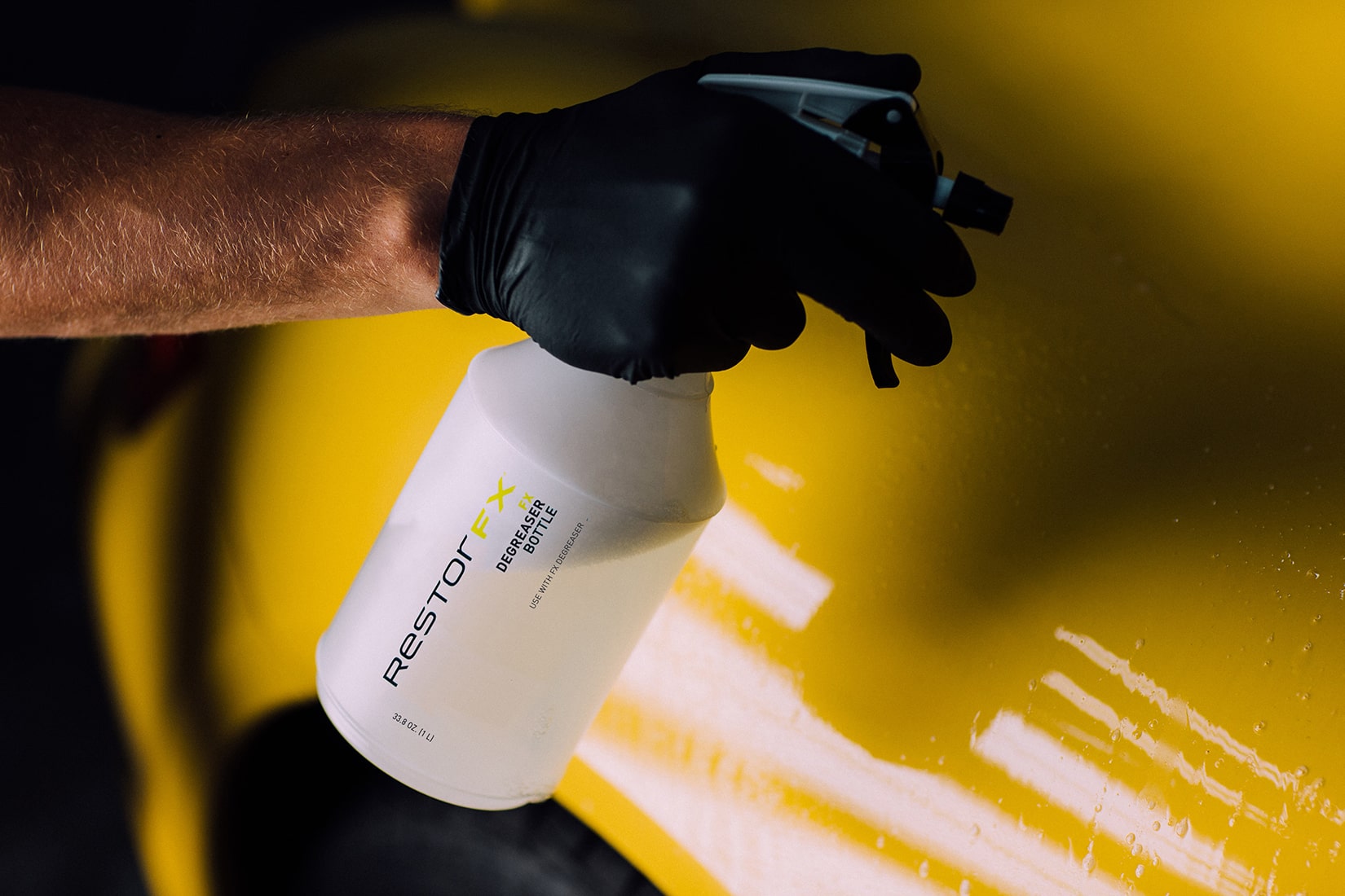 A gloved RestorFX technician degreasing the surface of a yellow sports car by spraying it with FX Degreaser