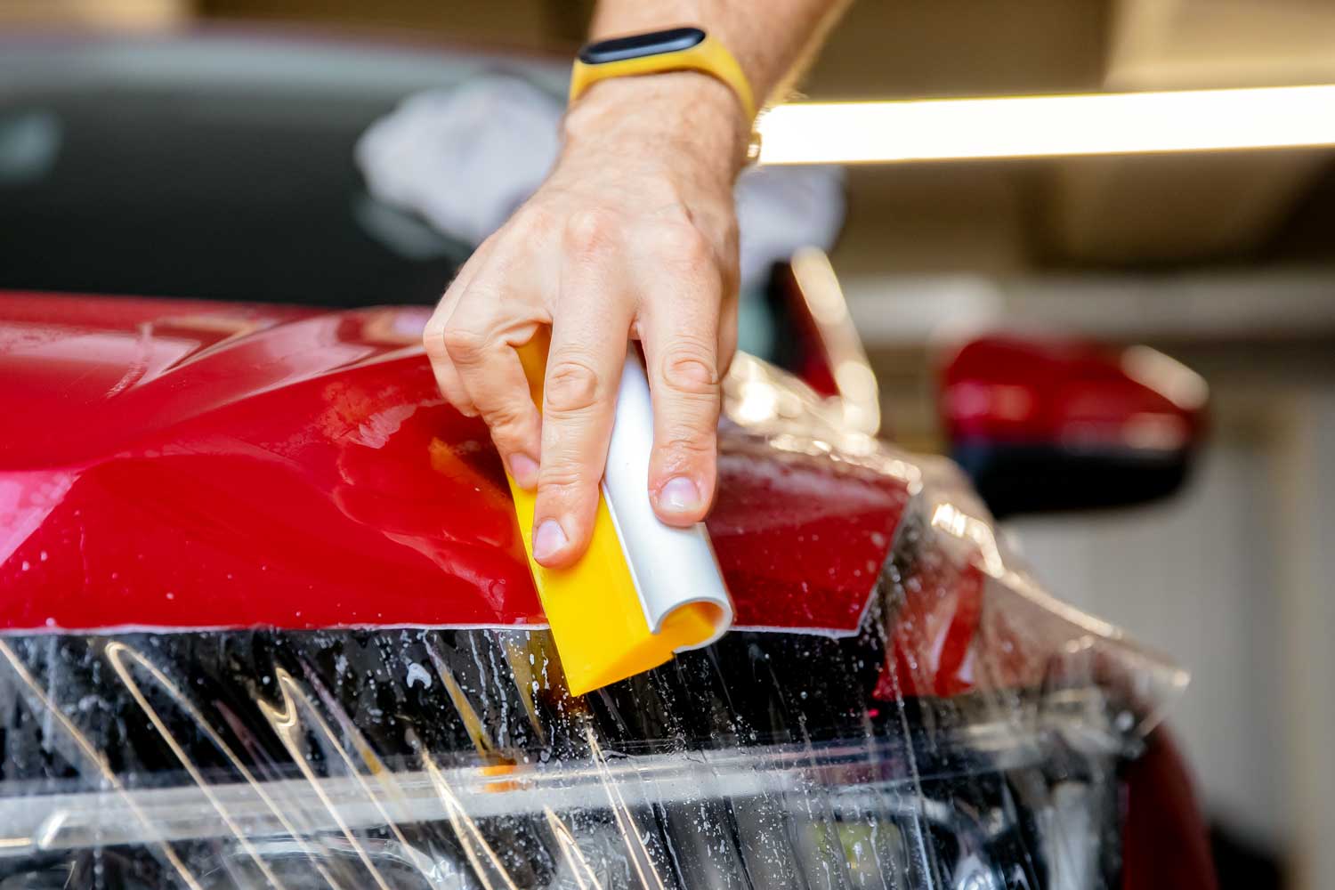 Technician applying clear protection film to the hood of a red car using a yellow squeegee